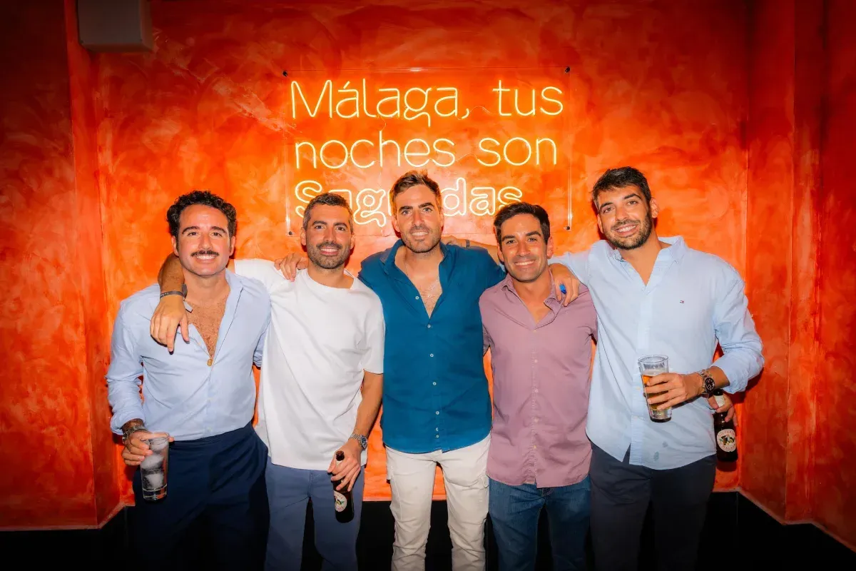 Cinco hombres posando juntos delante de un letrero de neón naranja que dice: 