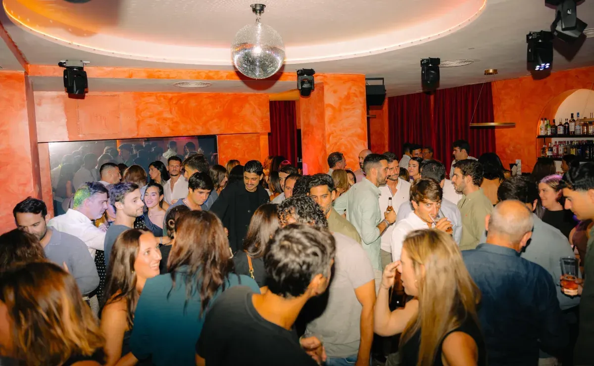 Multitud bailando en una discoteca con paredes naranjas, bola de discoteca y barra.