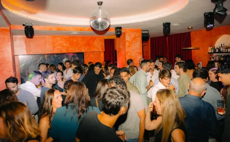 Multitud bailando en una discoteca con paredes naranjas, bola de discoteca y barra.