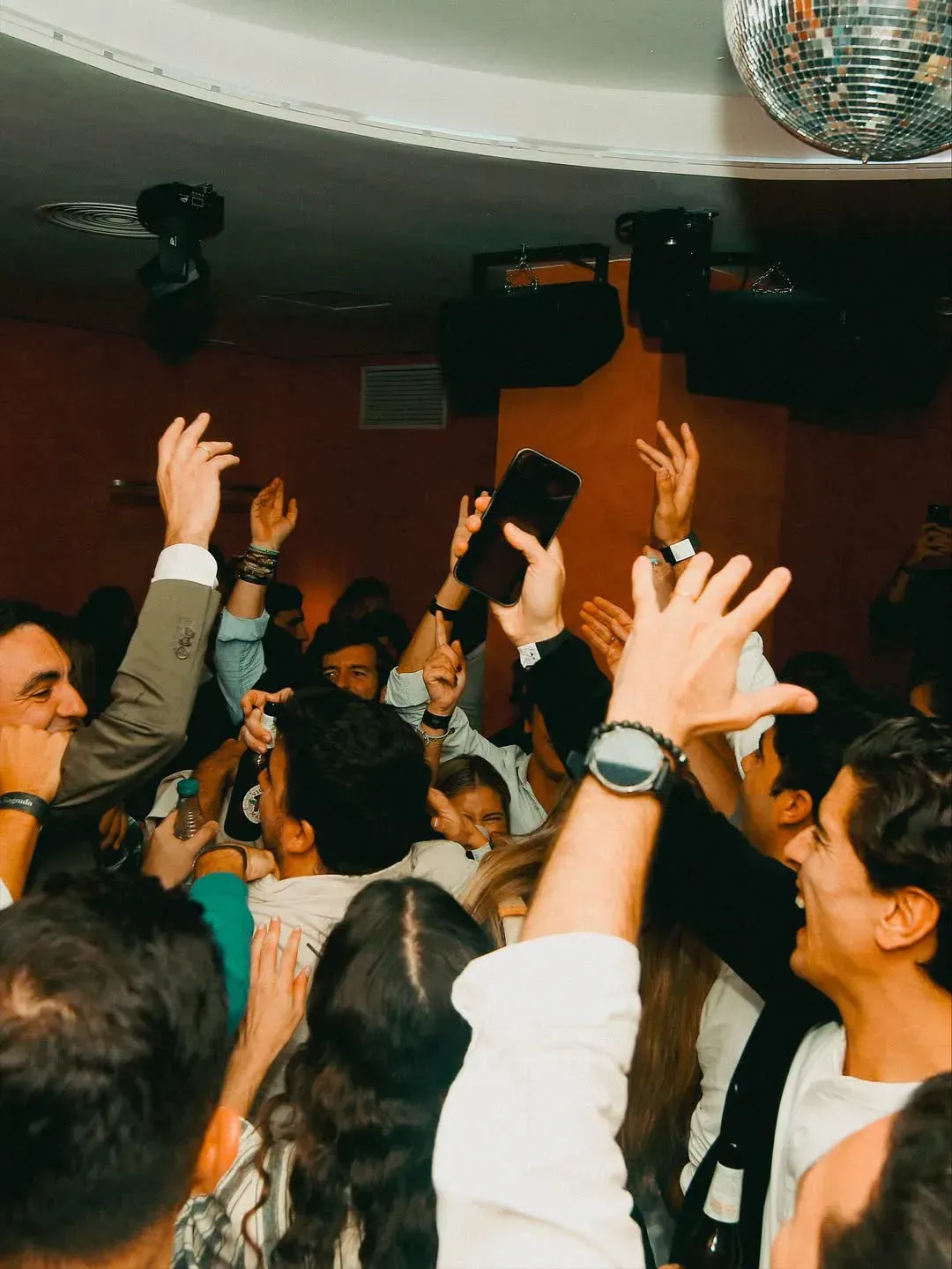 Gente bailando en un club, con los brazos en alto. Se ven la bola de discoteca, los altavoces y las paredes rojas.