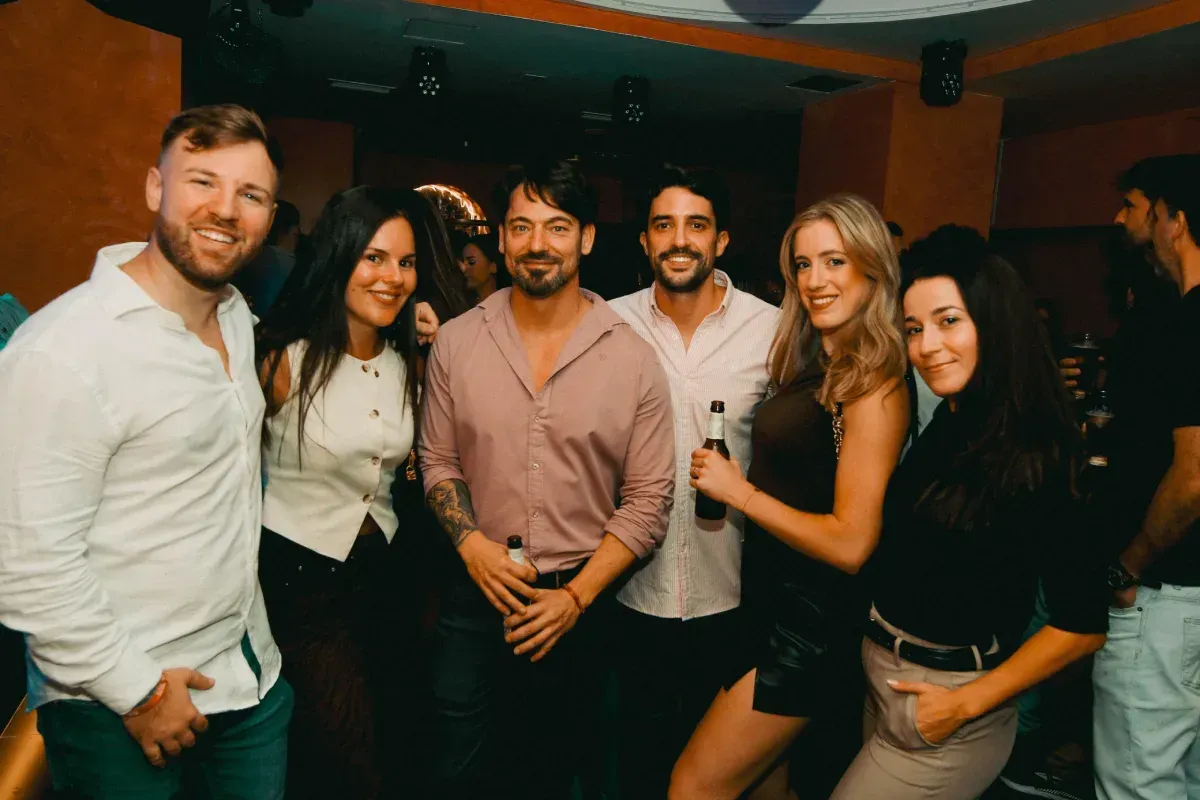 Grupo de personas sonriendo en una discoteca, algunas con bebidas en la mano. Iluminación cálida y fondo oscuro.