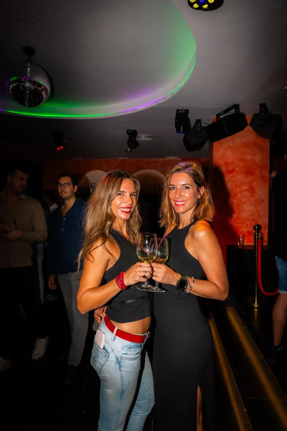 Dos mujeres en una discoteca, con bebidas en la mano. Una lleva vaqueros, la otra un vestido negro.