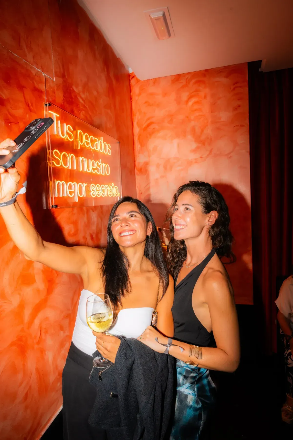 Dos mujeres se toman una selfie en una habitación naranja muy iluminada. Una sostiene un vaso