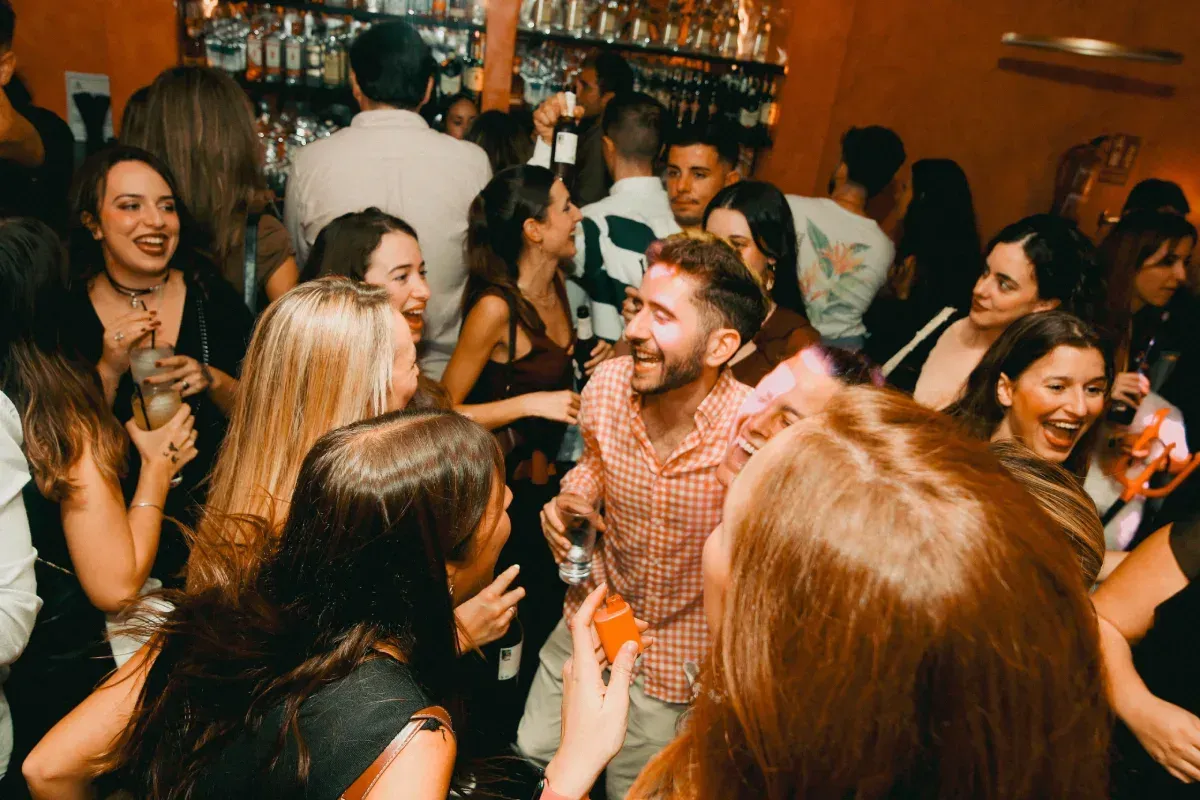 Gente socializando, riendo y bebiendo cócteles en un bar abarrotado con una barra al fondo.
