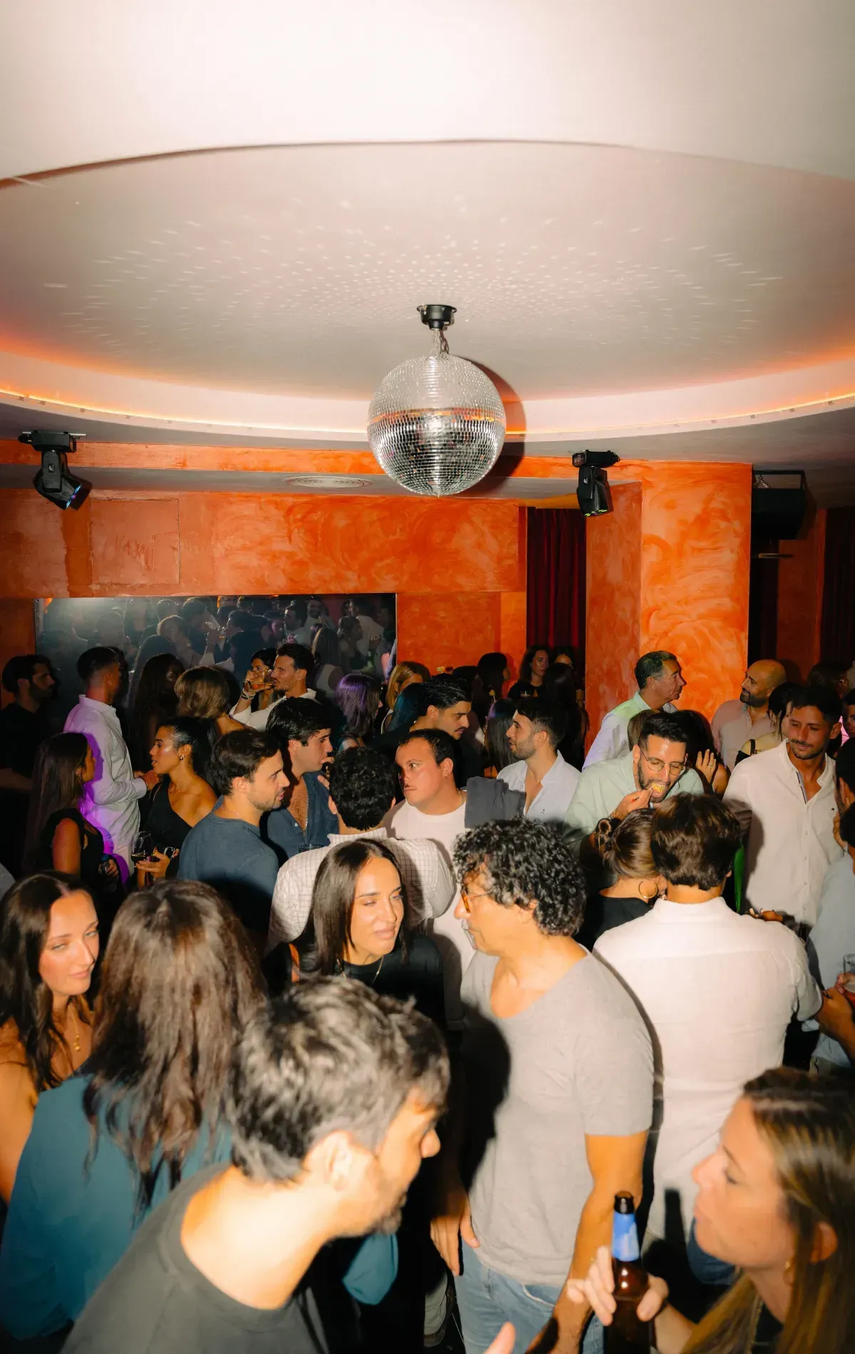 Discoteca abarrotada, gente charlando, bolas de discoteca colgando. Iluminación cálida, paredes naranjas, vestimenta variada.