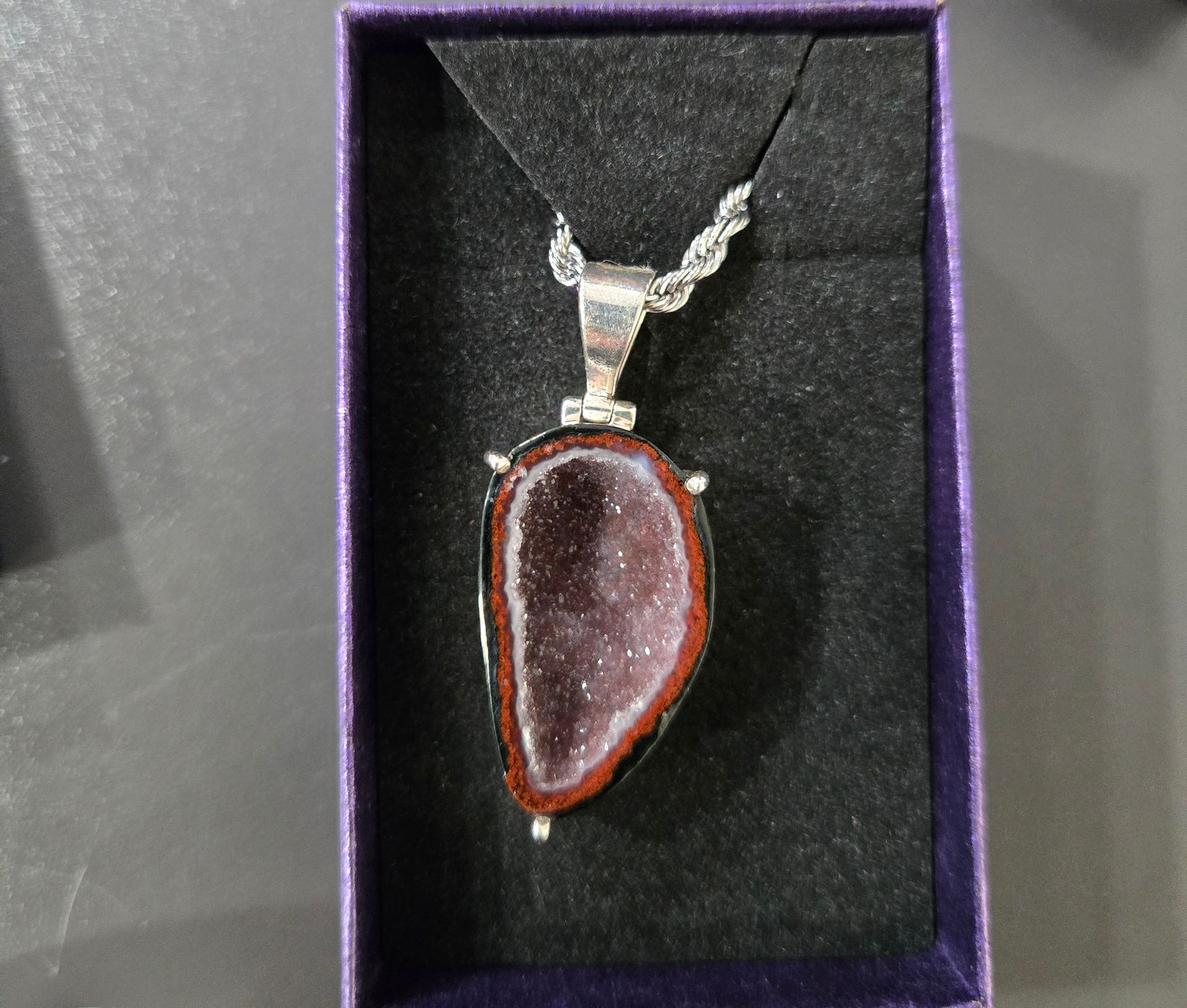 Pendentif en forme de larme violette sur une chaîne en argent, présenté dans un écrin en velours.