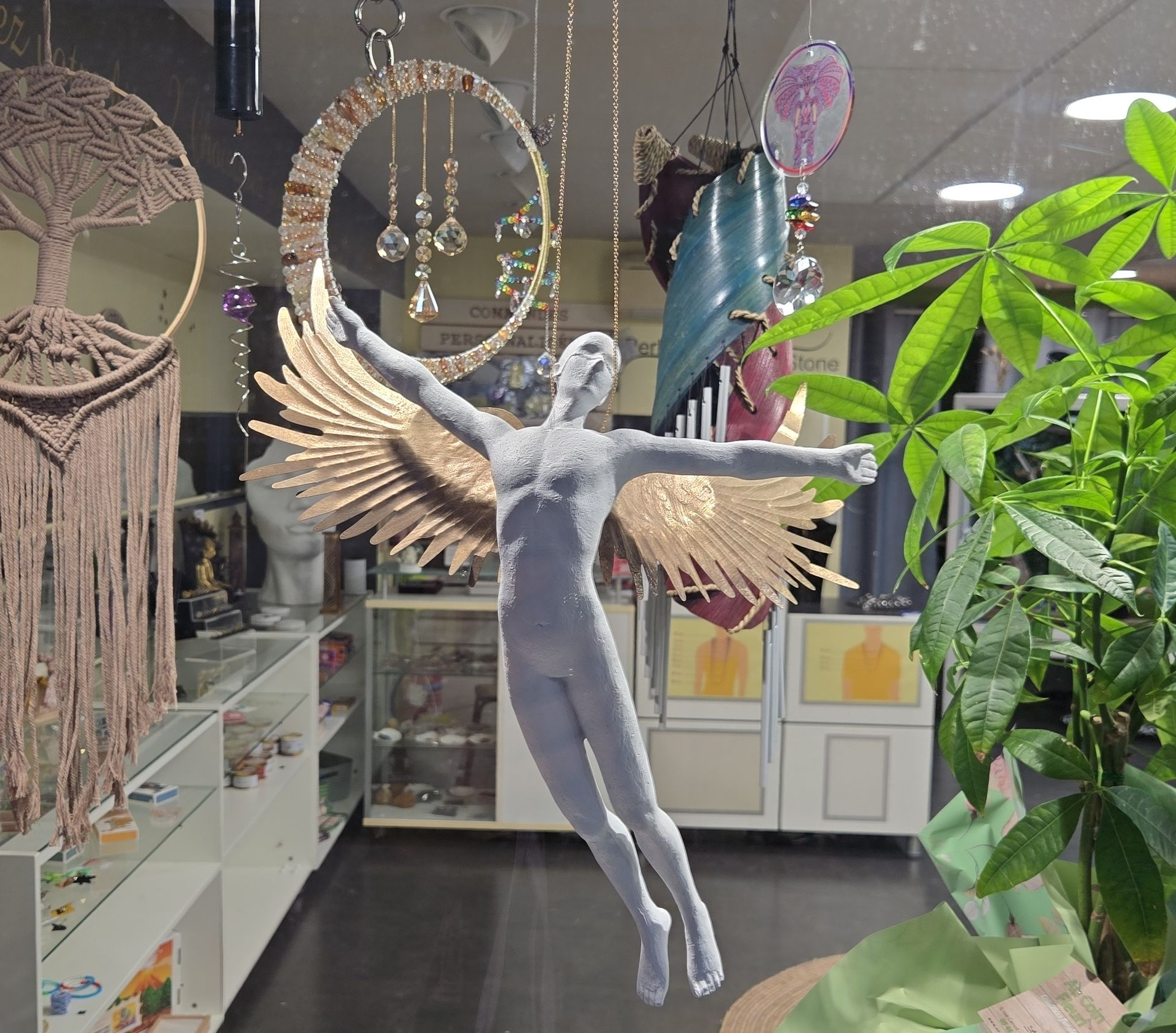 Une fée ornementale aux ailes argentées est suspendue dans une boutique remplie de plantes, avec des étagères et des éléments de décoration en arrière-plan.