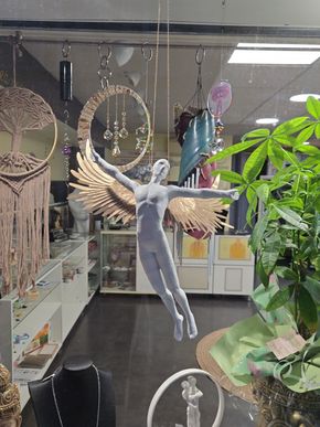 Une figurine d'ange est suspendue parmi des plantes et des objets artisanaux dans une vitrine.
