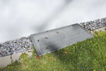 Rechteckiger Metall-Geräteschacht neben einem grasbewachsenen Wegrand und einem mit Steinen gesäumten Pfad