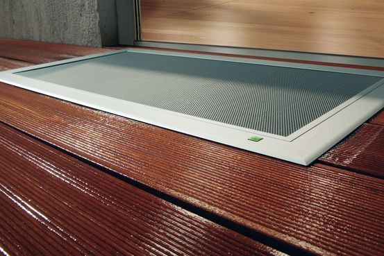 Eine weiß gerahmte Lichtschachtabdeckung auf einer rötlichen Holzterrasse vor einer holzgetäfelten Wand