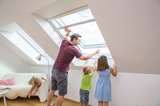 Eine Familie öffnet die Dachfenster in einem hellen Dachzimmer; ein Kind sitzt in der Nähe auf einem Sofa.