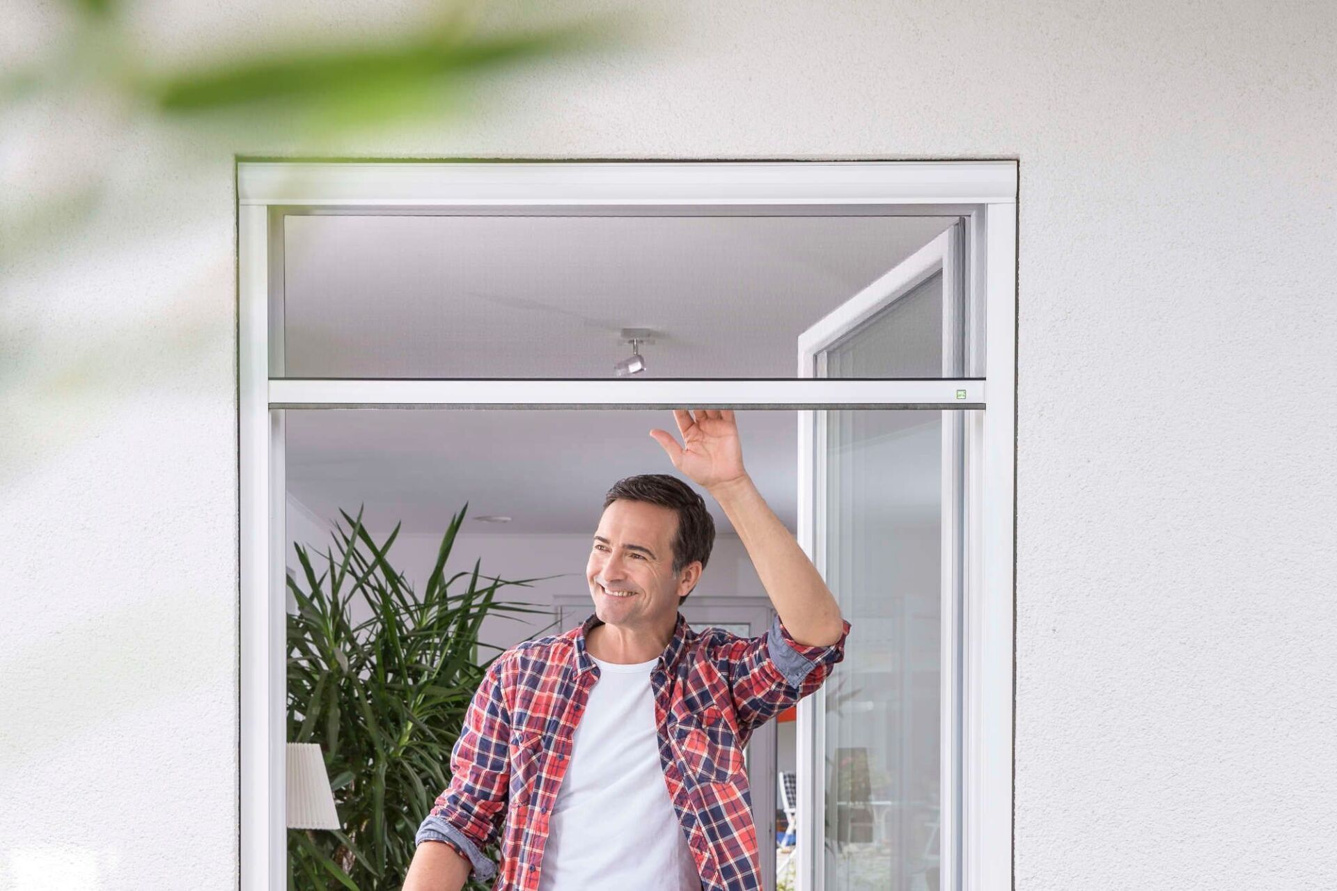 Ein Mann in einem karierten Hemd winkt aus einem offenen Fenster Ein Mann in einem karierten Hemd winkt aus einem offenen Fenster neben einer Topfpflanze.