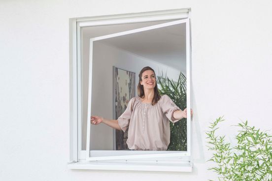 Eine Frau öffnet ein weißes Fenster Eine Frau öffnet ein weißes Fenster in einem hellen Zimmer; im Hintergrund sind eine Pflanze und ein Wandbild zu sehen.