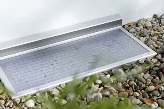 Ein silberner Metallablaufrost neben Kieselsteinen, davor ein flacher, erhöhter Rand und verschwommenes Grün.