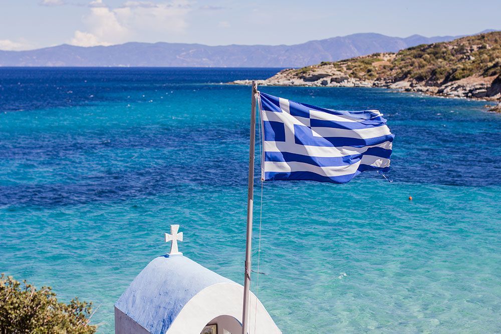 Vor einer kleinen Kirche am Strand weht eine griechische Flagge.