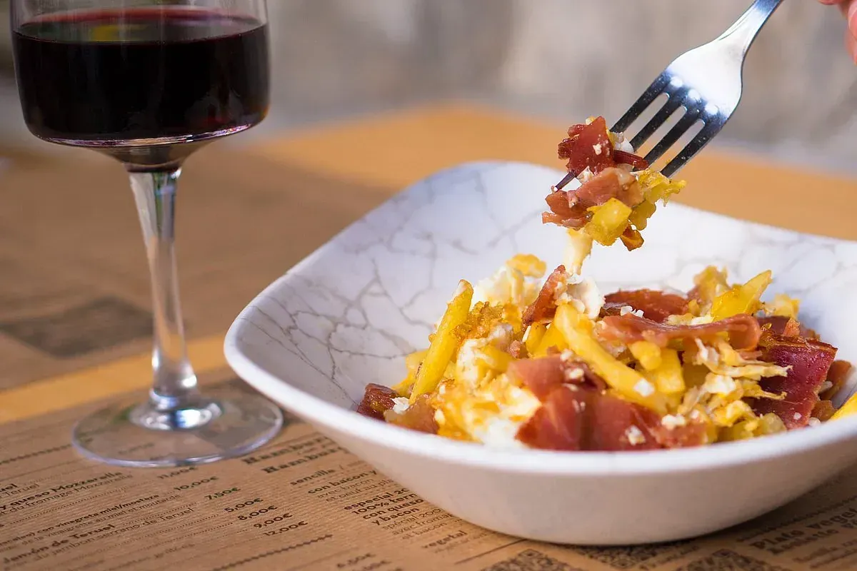 Un tazón de papas fritas con tocino y queso, una comida para levantar con tenedor y una copa de vino tinto.