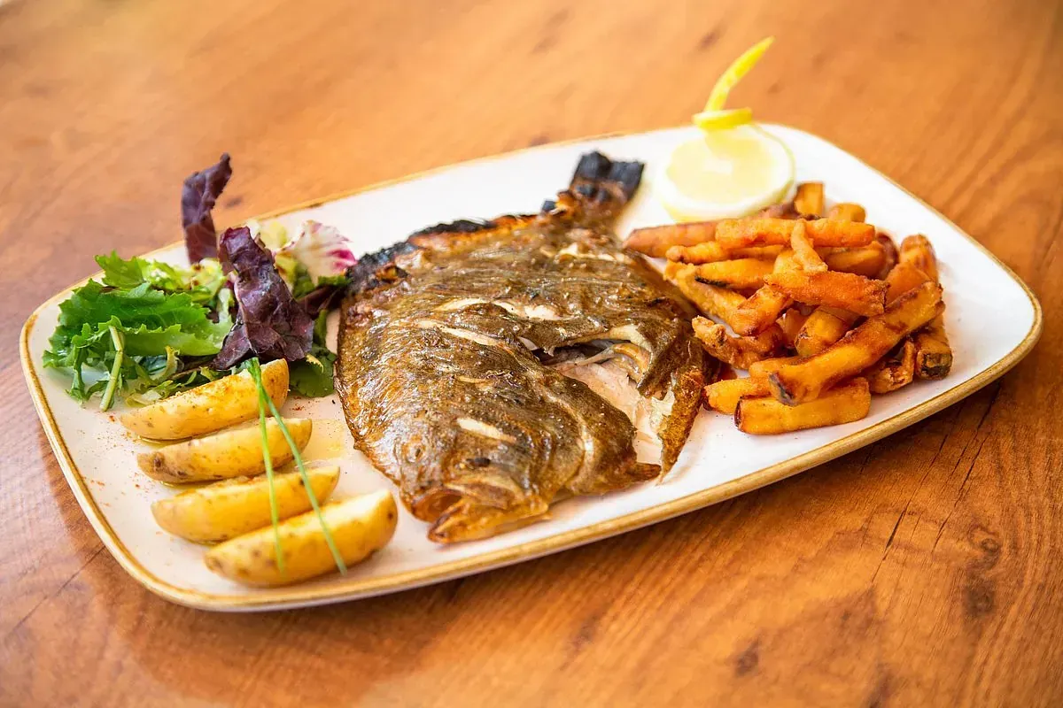 Pescado a la plancha, patatas fritas y gajos de patata en un plato rectangular con una rodaja de limón y verduras, dispuesto sobre una mesa de madera.
