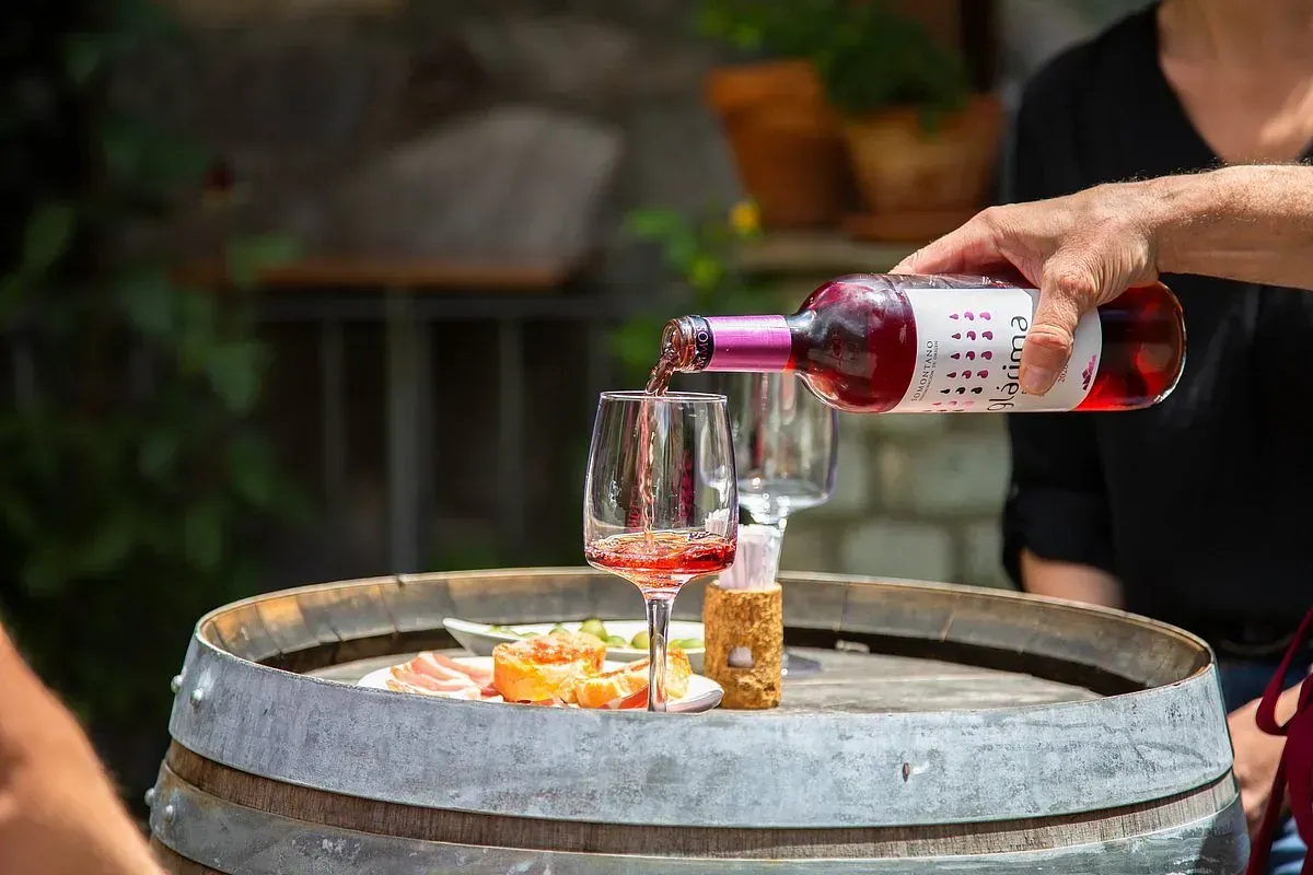 Vino rosado servido en una copa sobre un barril de madera, con comida y una persona al fondo.