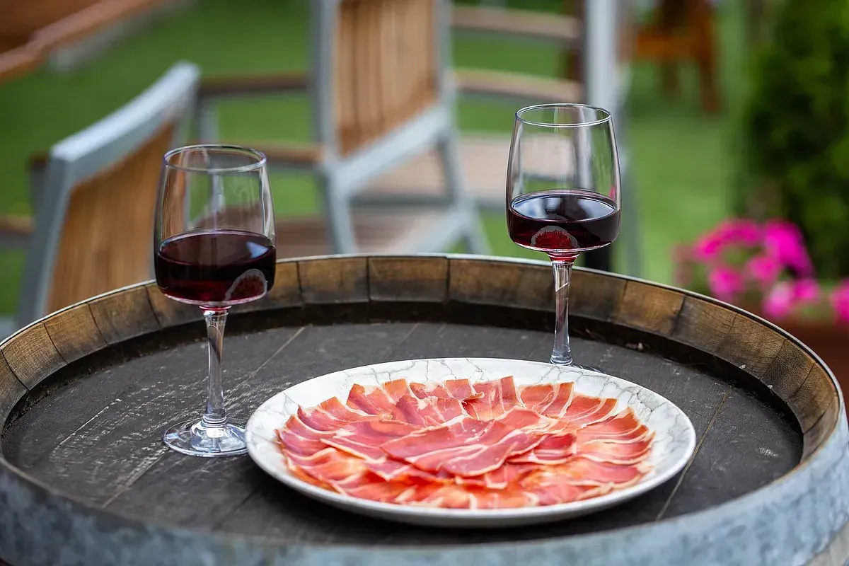 Dos copas de vino tinto y un plato de carne en rodajas sobre un barril de madera al aire libre.