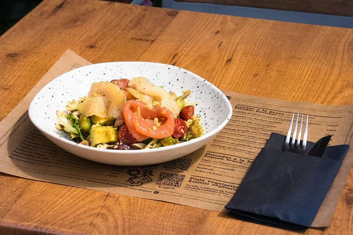 Ensalada en un bol moteado sobre una mesa de madera, junto a un tenedor y una servilleta negra doblada.