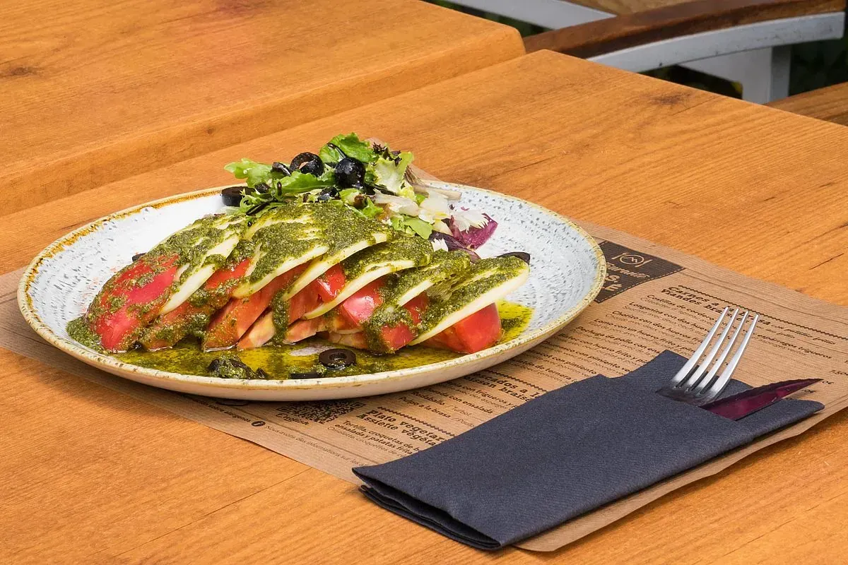 Ensalada caprese con salsa pesto, en un plato con hojas verdes. Tenedor y cuchillo sobre una servilleta, mesa de madera.