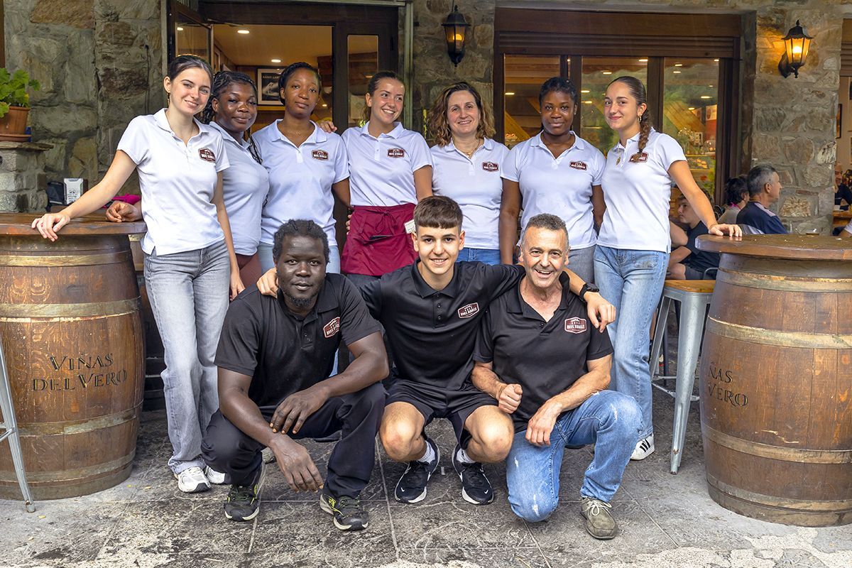 Grupo posando frente a un restaurante rústico, con empleados vestidos con camisas blancas y tres personas arrodilladas delante.