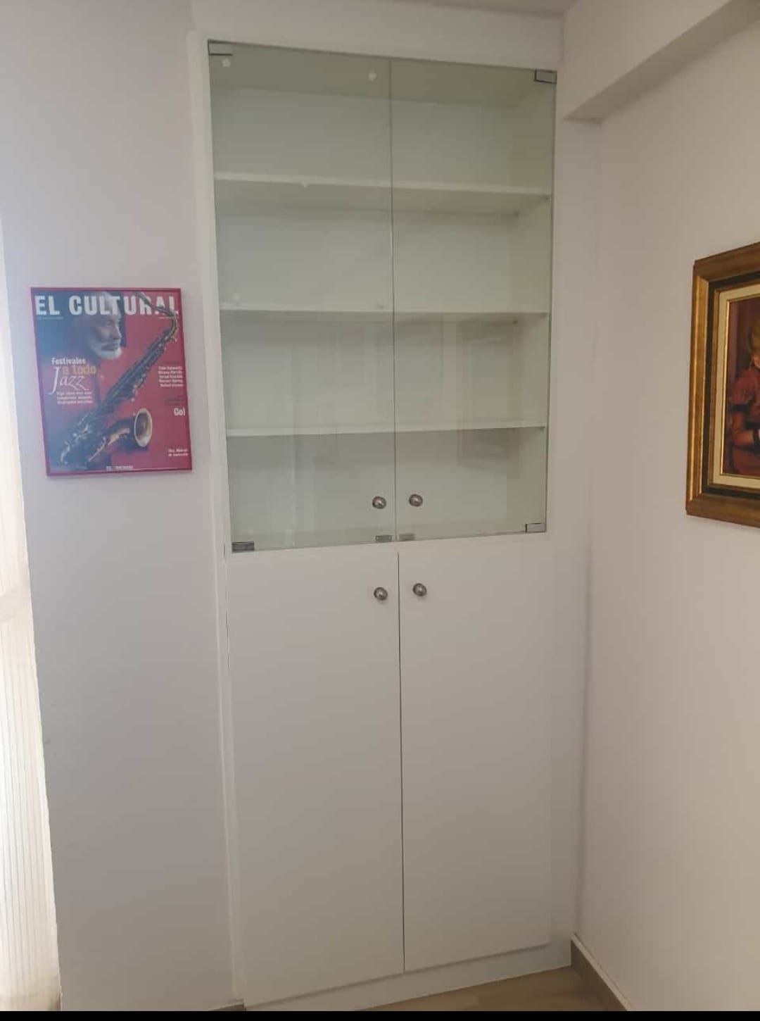Mueble empotrado blanco con estantes superiores con puertas de cristal y puertas de almacenaje inferiores, junto a una pared con obras de arte.