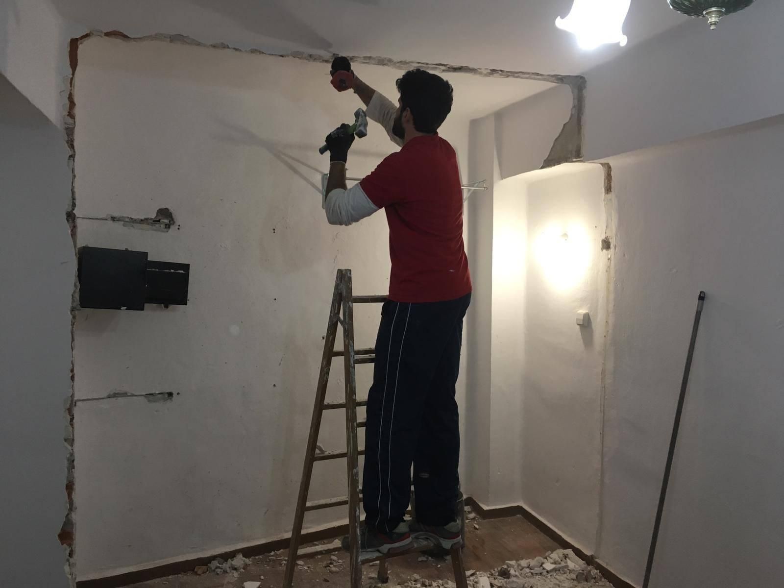 Un hombre con camisa roja está parado en una escalera y usa una herramienta para quitar una sección de una pared blanca.