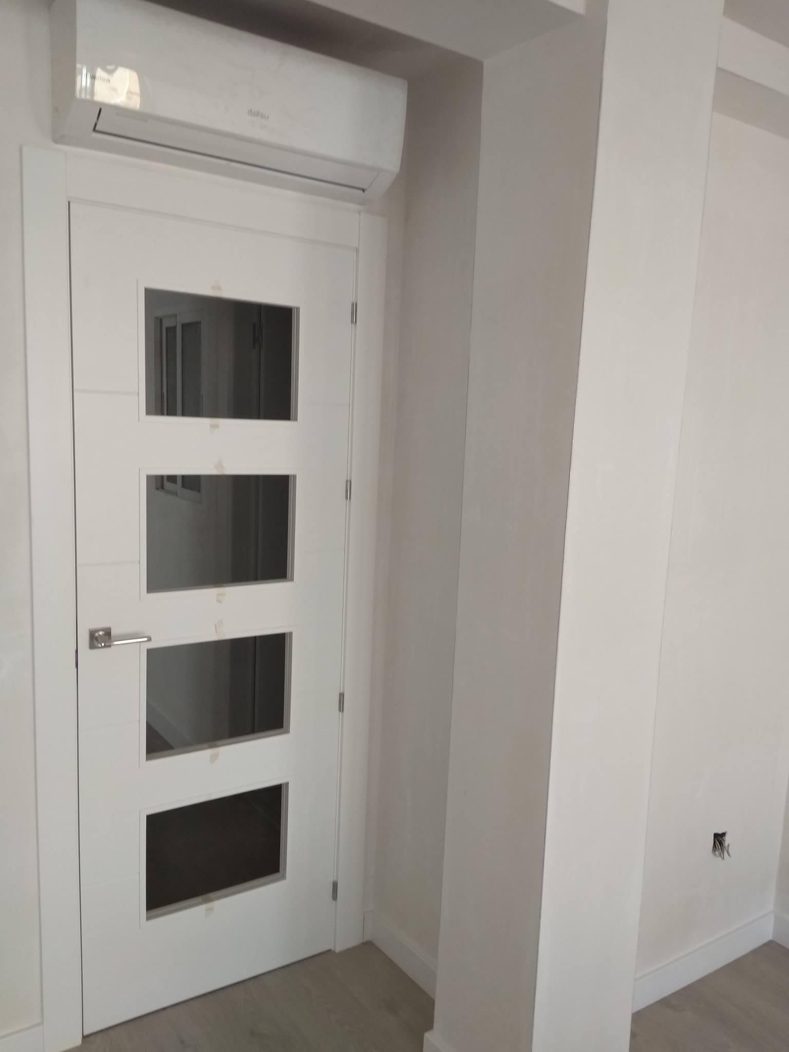 Puerta blanca con cuatro paneles de vidrio, con aire acondicionado encima. En una habitación con paredes blancas.