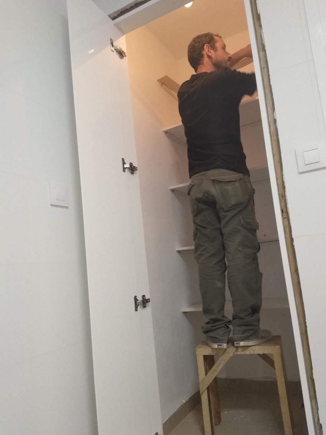 Hombre de pie sobre un pequeño taburete, instalando estantes en un armario blanco.