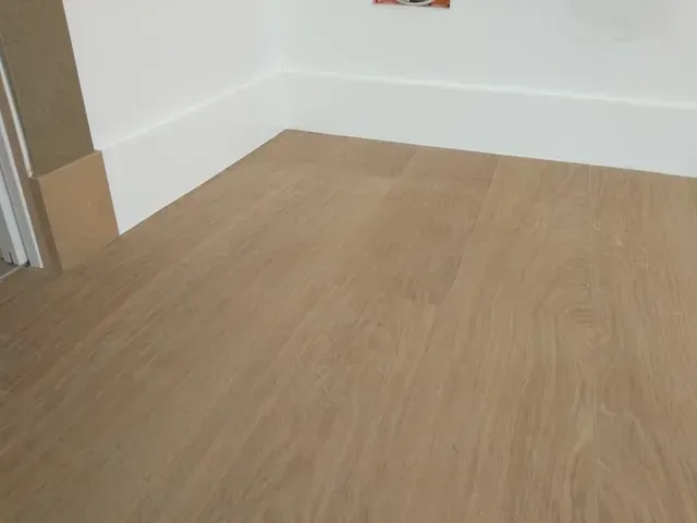 Piso de madera que se junta con zócalo blanco y pared; toma de corriente visible.