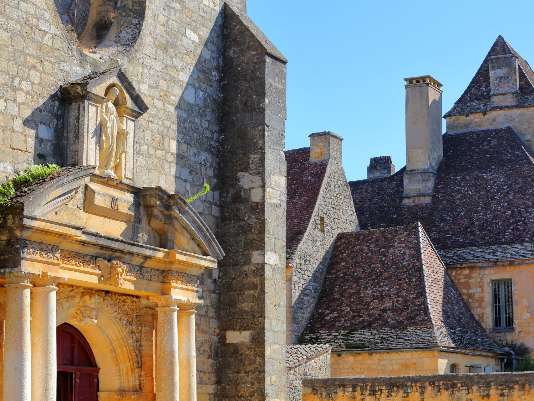 Façade d'église en pierre avec une statue et des toits de tuiles rouges se détachant sur un ciel bleu clair.