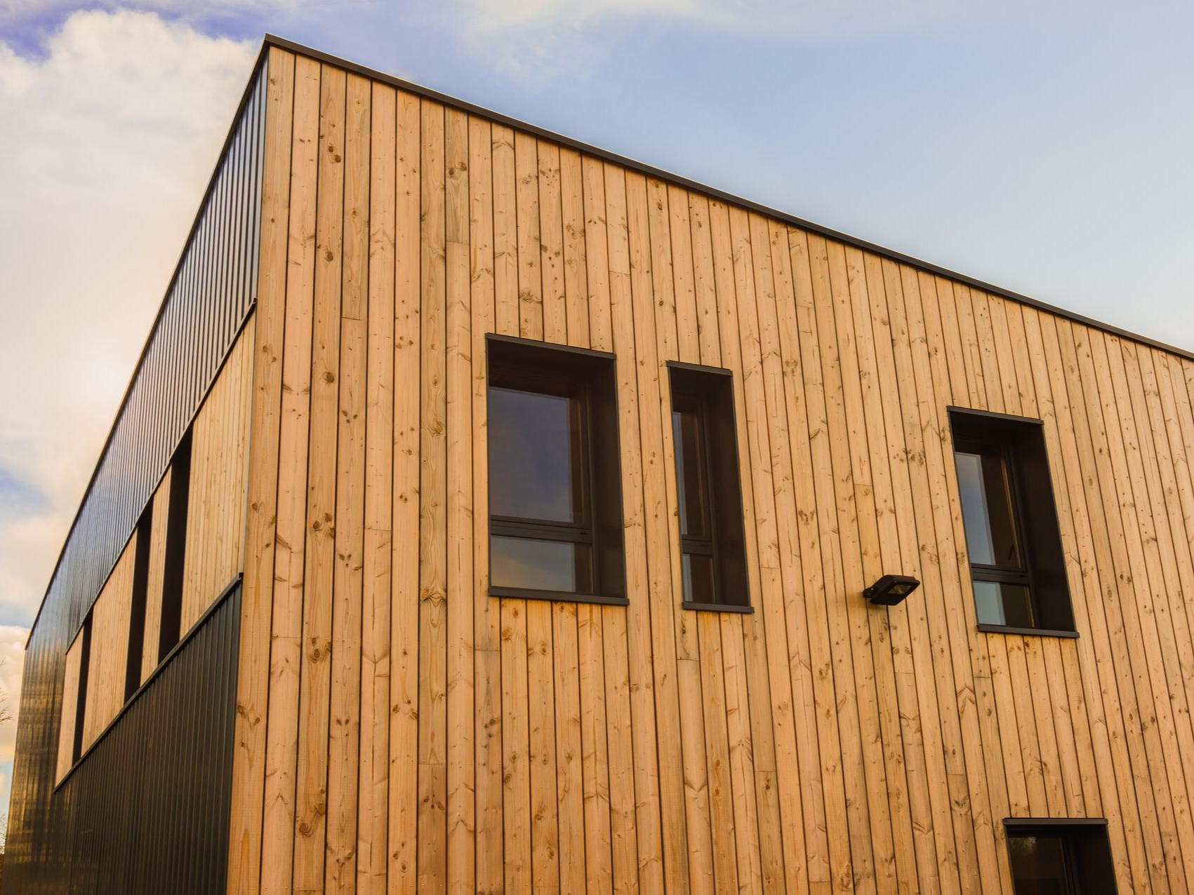 Façade d'un bâtiment moderne avec bardage vertical en bois et fenêtres à cadre sombre sur un ciel partiellement nuageux.