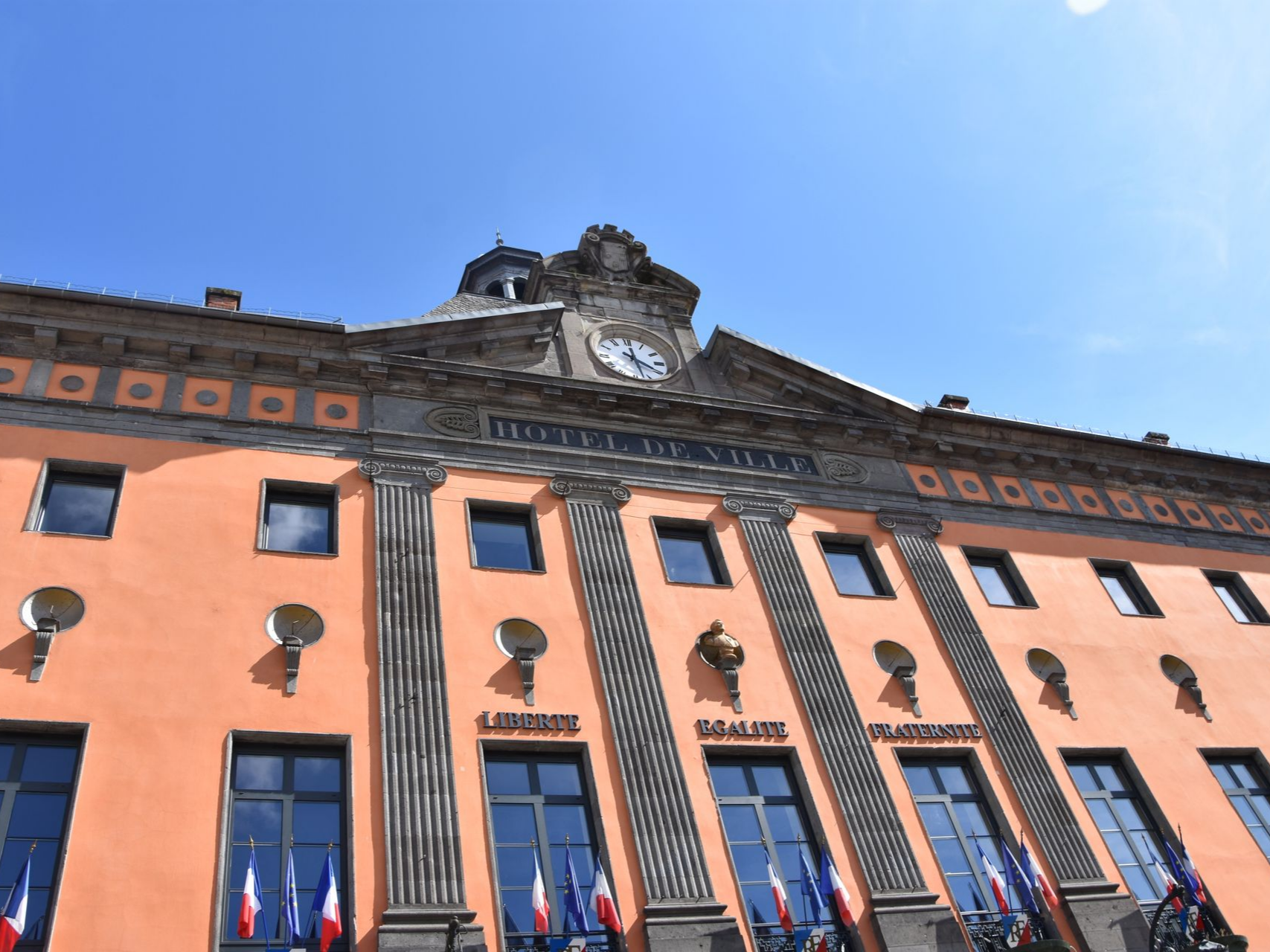 Bâtiment de la mairie couleur saumon avec horloge, colonnes et drapeaux français, sous un ciel bleu.
