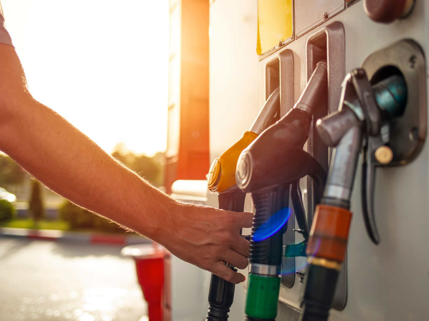 La main d'une personne se tend vers le pistolet bleu d'une pompe à essence dans une station-service, avec la lumière vive du soleil en arrière-plan.