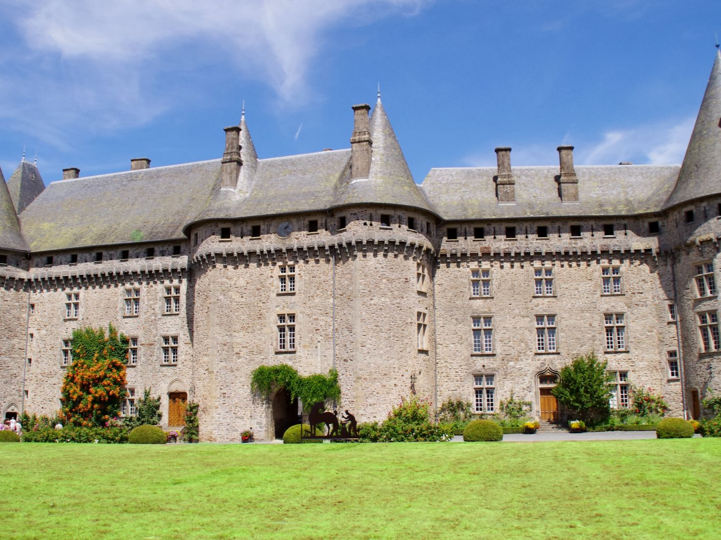 Grand château en pierre avec plusieurs tours, toit gris et pelouse verte par une journée ensoleillée.
