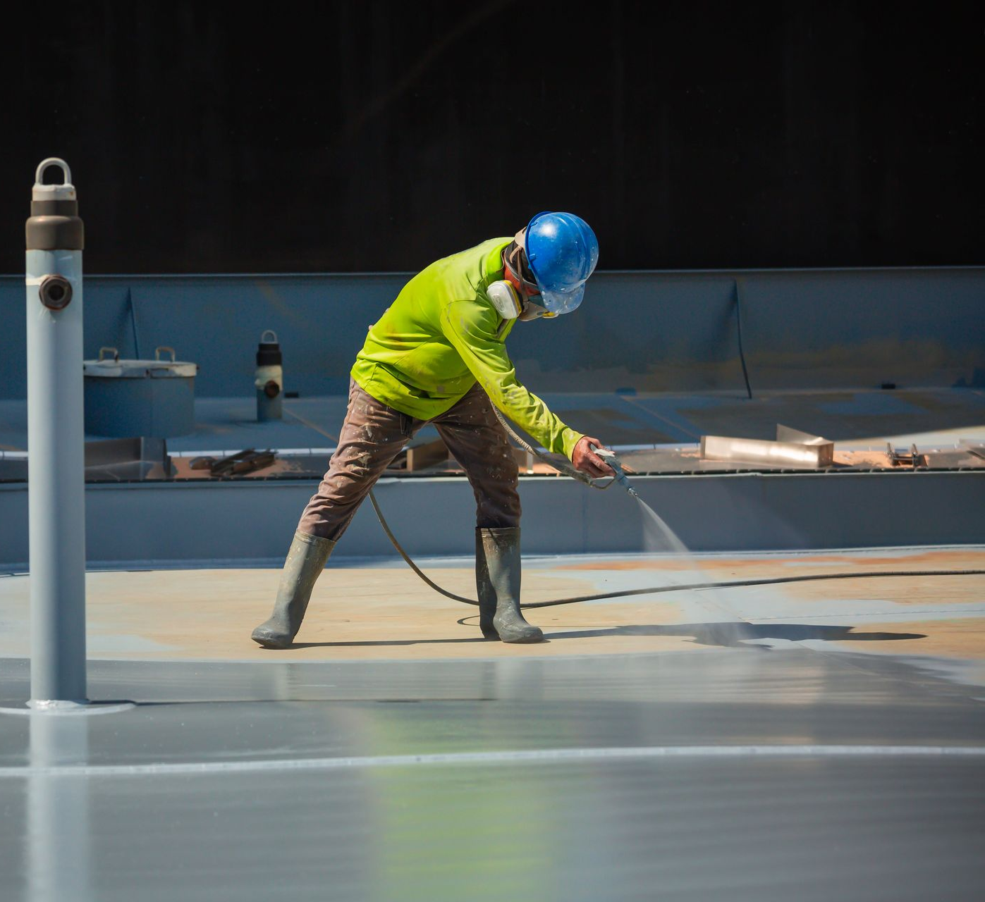 Un ouvrier portant un casque bleu et une chemise fluo vaporise de la peinture sur un sol gris sur un chantier de construction.