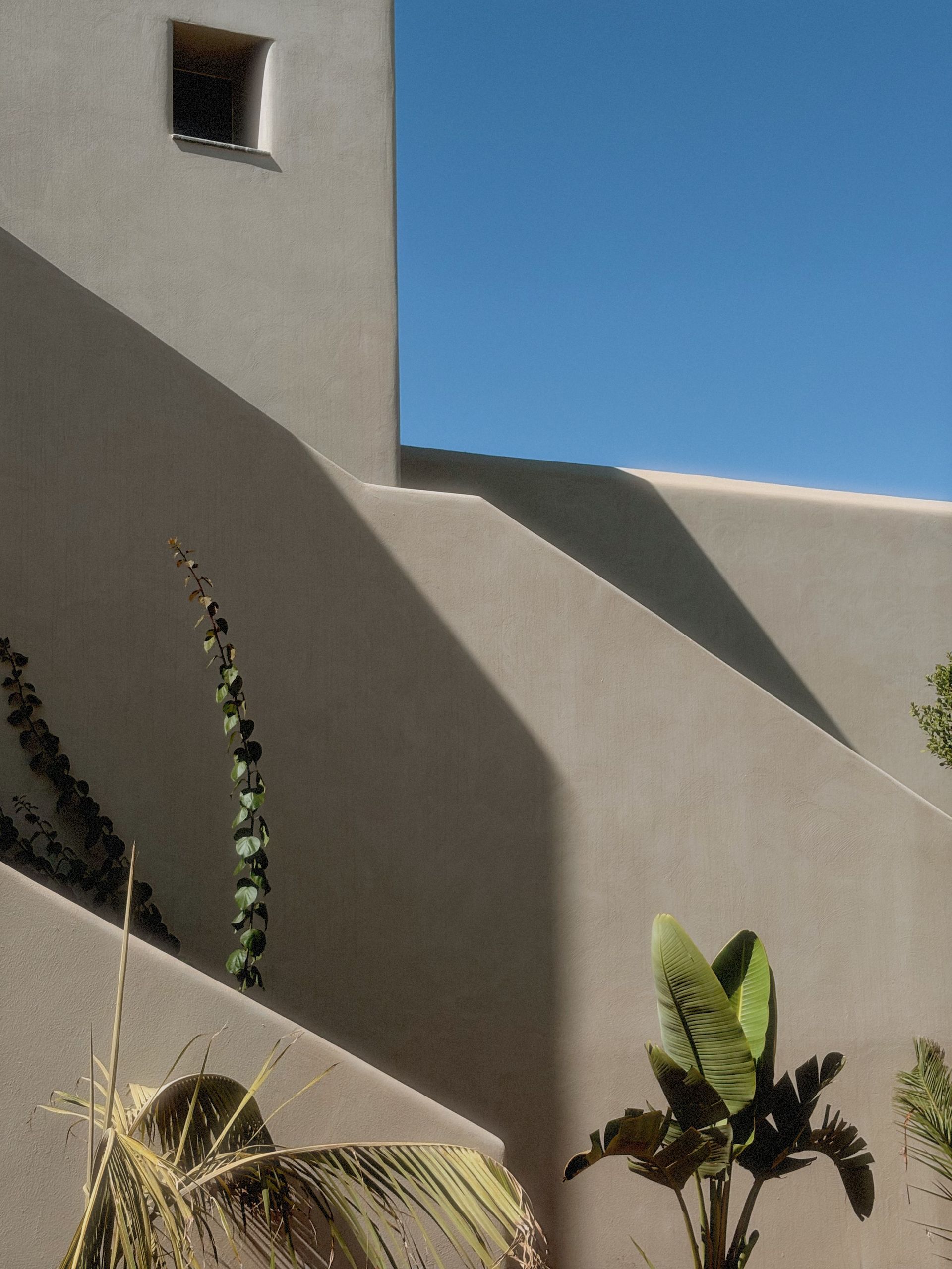 Edificio de estuco beige con sombras y plantas contra un cielo azul.