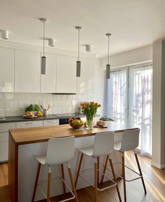 Cocina moderna con armarios blancos, isla de madera con tres taburetes y mucha luz natural.