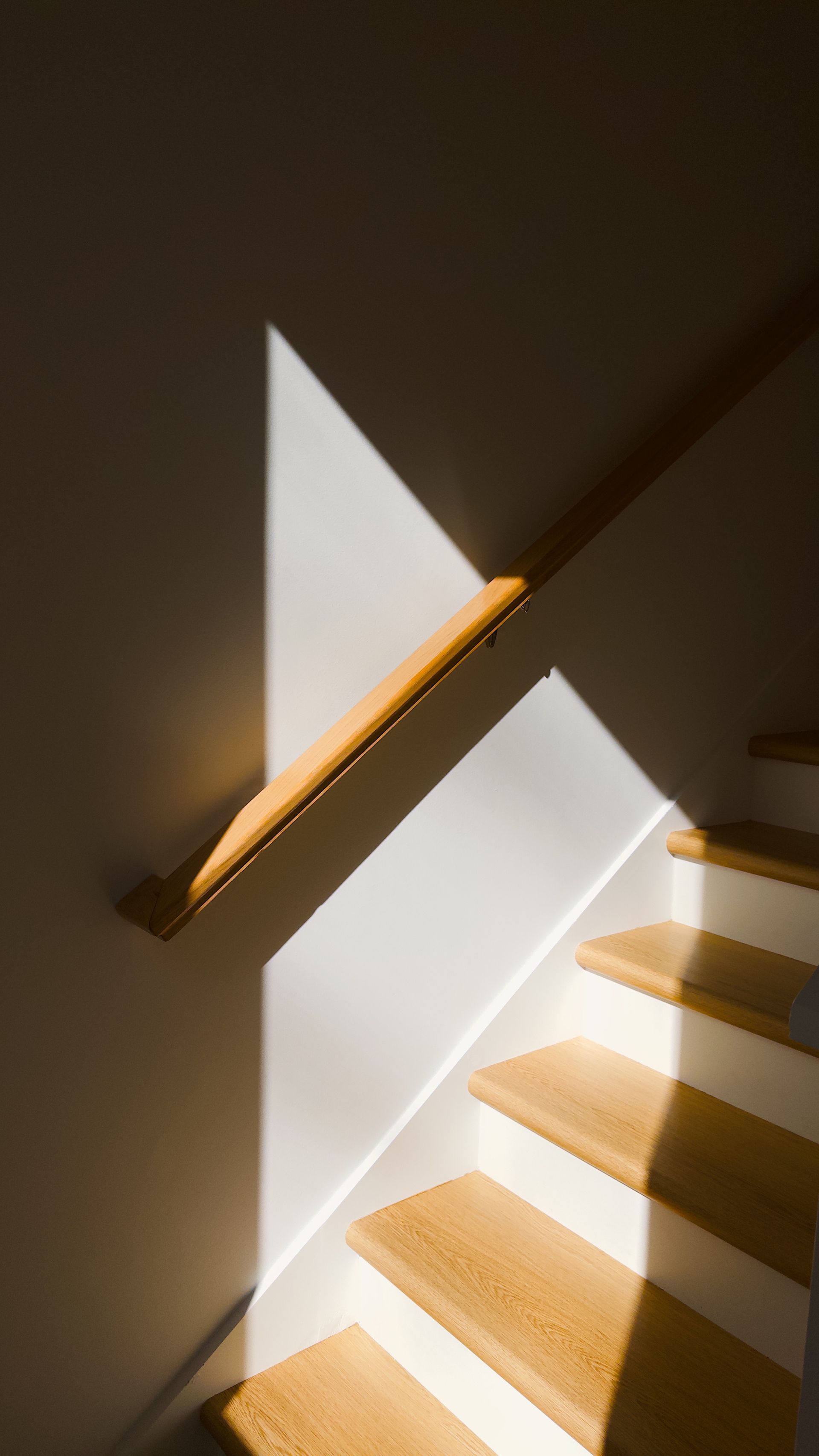 Escalera con peldaños y barandilla de madera, iluminada por un haz triangular de luz solar sobre una pared blanca.