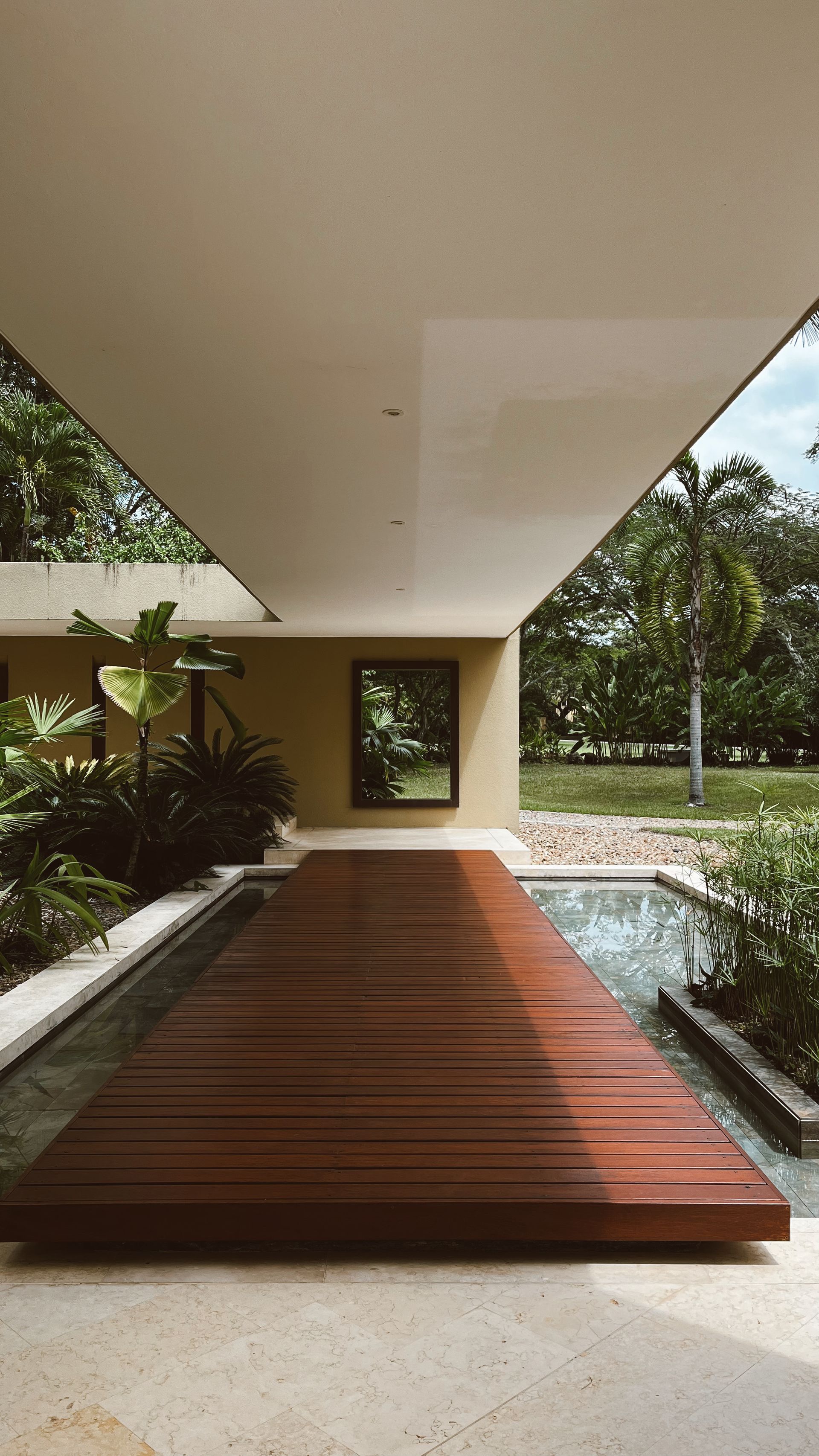 Una pasarela de madera sobre el agua conduce a la entrada del edificio; rodeada de plantas y exuberante vegetación.