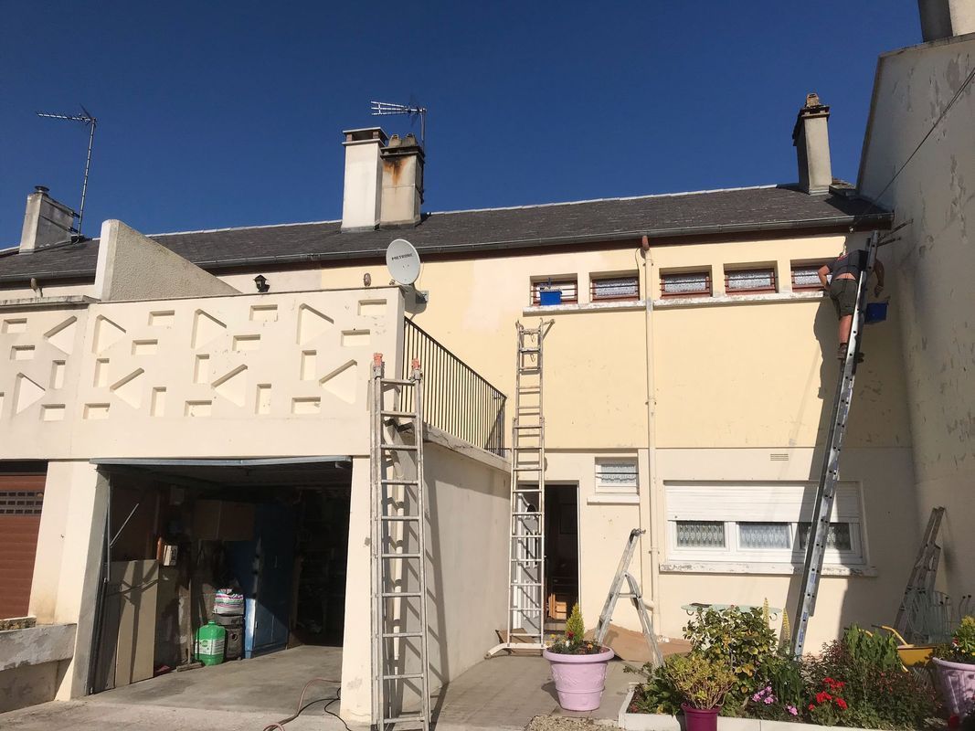 Maison beige à deux étages avec garage, cheminées sur le toit et fleurs en pot.