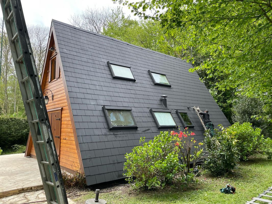 Chalet moderne noir en forme de A avec puits de lumière, situé au milieu des arbres et des arbustes.
