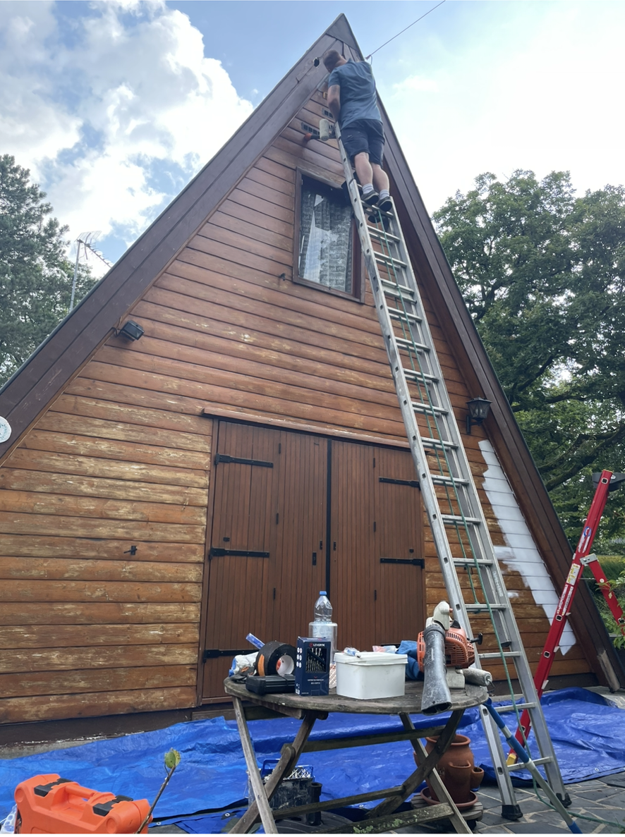 Extérieur d'une cabane en A avec des ouvriers sur une échelle près du toit, des outils et des fournitures sur une bâche bleue en dessous.