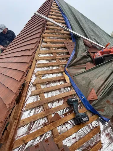 Toiture en réparation, avec chevrons en bois apparents, solins métalliques et un ouvrier sur la gauche.