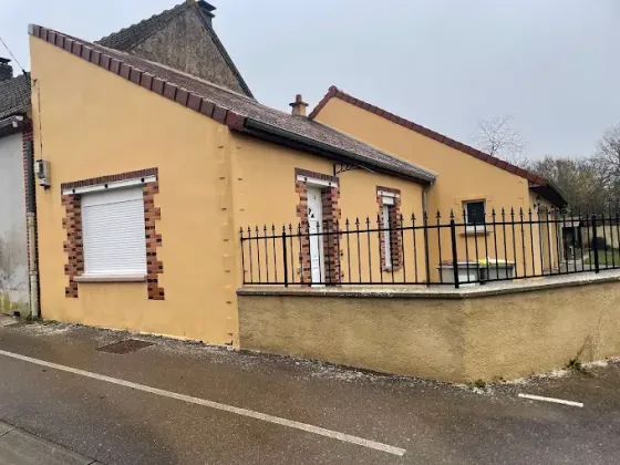 Maison jaune aux fenêtres à volets et à la clôture métallique noire, située dans une rue tranquille.