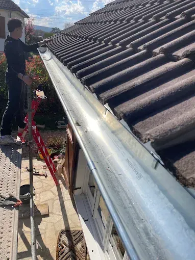 Un ouvrier inspecte une gouttière argentée le long d'un toit de tuiles par une journée ensoleillée.