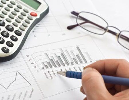 Mão segurando uma caneta sobre gráficos com calculadora e óculos em uma mesa