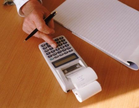 Mão apontando para uma calculadora ao lado de um bloco de notas em uma mesa de madeira com uma impressora de recibos.
