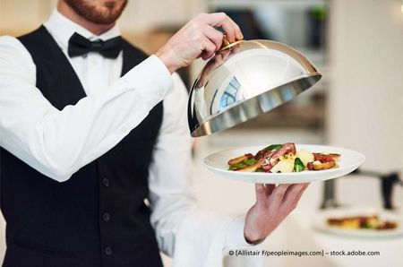 Ein Kellner hält eine Speiseglocke über einen Teller mit Essen.